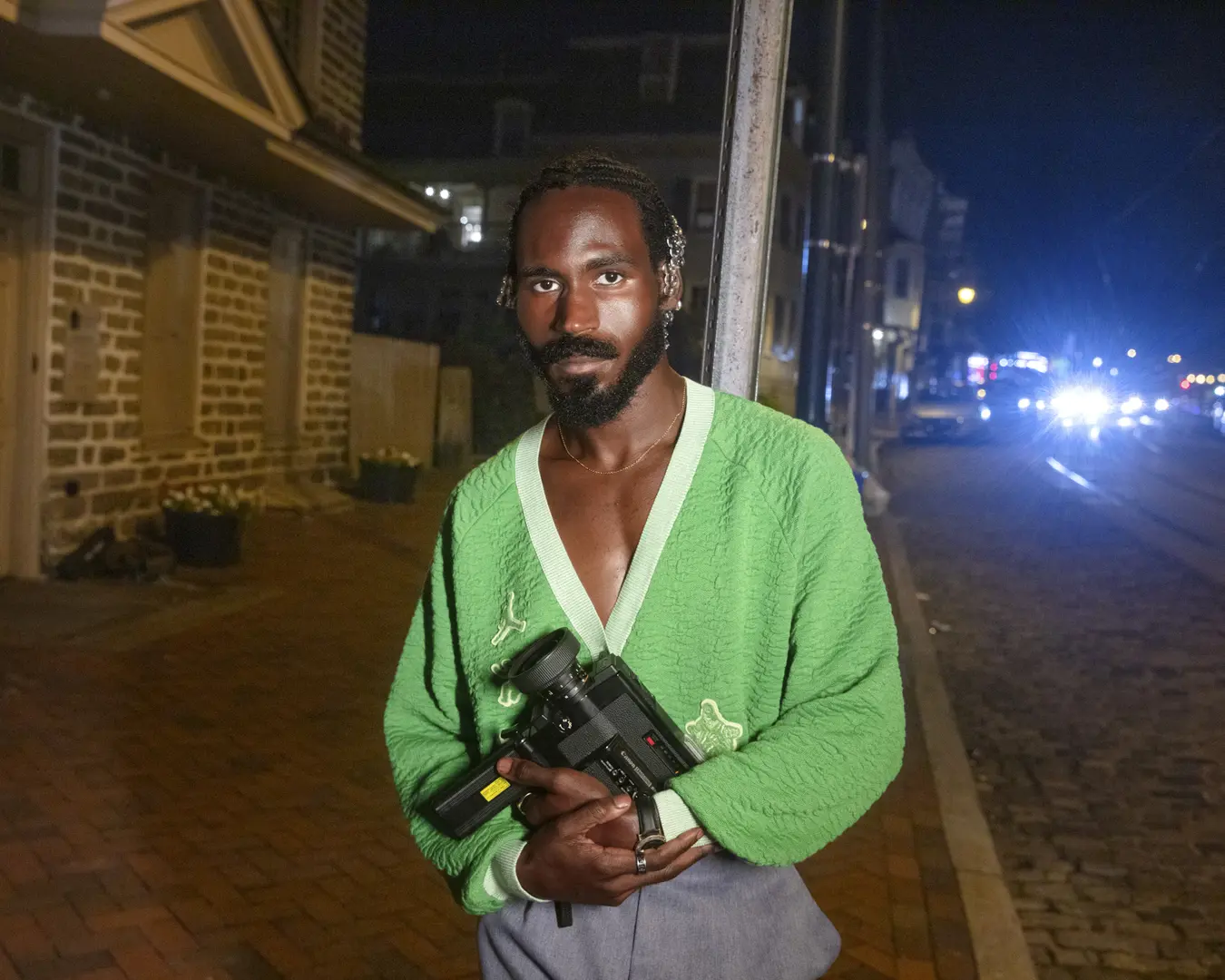 A portrait of filmmaker Vernon Jordan. He stands outside at night holding his video camera. He is a Black man with facial hair wearing a green v-neck cardigan.