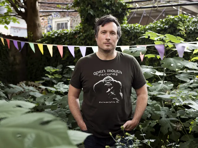 Bill Nace stands amidst waist-high plants with a string of decorative flags just behind him. An ivy-covered wall and electrical wires are visible in the background. He has light skin and short brown hair and wears a black record label t-shirt.