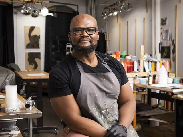 Didier sits on a stool in his studio. He has brown skin, a bald head, and black facial hair. He wears a black t-shirt, pink pants, a gray paint-spattered apron, black latex gloves, and dark-framed glasses.