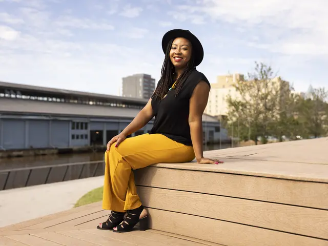 Dinita Clark smiles sitting on the steps of Philadelphia’s Race St. Pier. She has warm-toned med-dark skin and wears her long brown hair in tight dreads. She wears bright yellow pants, a black sleeveless top, black heeled sandals, and a black hat. 