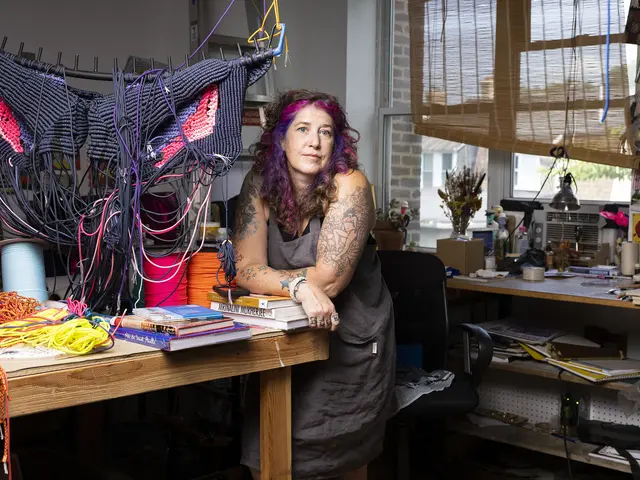 Jesse Harrod stands leaning on their desk in their studio. They have light skin, visible tattoos, and long curly hair dyed pink and purple around their face. A purple and pink artwork in progress hangs near their head with loose yarn hanging down. 