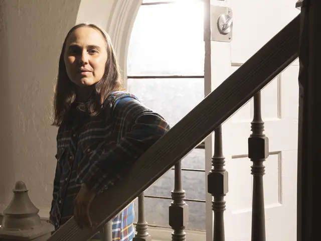 Julian Talamantez Brolaski stands on a staircase in front of an arched window, their face half in shadow. They have medium skin and long brown hair and wear a blue plaid shirt tucked into black denim, and a brown belt.