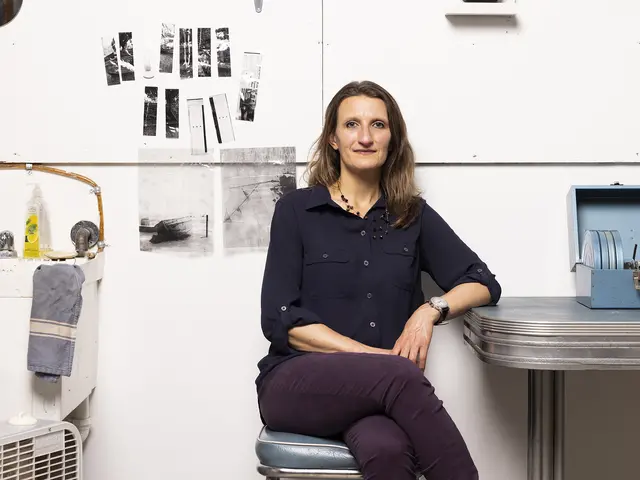 Lisa Marie Patzer sits in her studio against a white wall, her elbow leaning on a metal table. She has light skin, brown hair and wears a blue button down blouse and dark purple pants. 