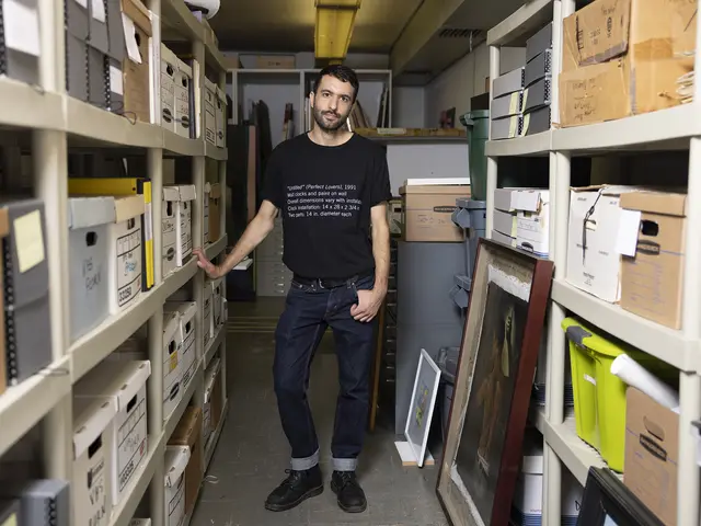 Rami stands in the midst of the archives of the William Way LGBT Community Center. They have medium-toned skin, short black hair, and short black facial hair. They wear a black t-shirt with placard text in white lettering, dark jeans, and black boots.