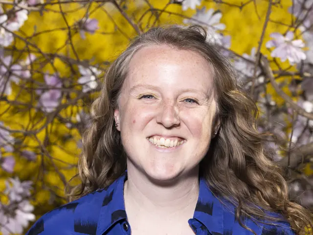 Emily grins joyfully at the camera. She has light skin, dark blonde hair, and blue eyes with gold eye shadow. She wears a bright blue shirt with black designs and has a nose ring. Blurry blossoming tress are visible behind her.