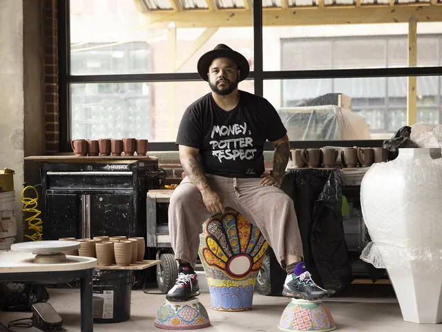 Roberto Lugo sits on one of his own large colorful ceramics in his studio. His feet are both resting on overturned bowls of his creation. He has medium skin, facial hair, & tattoos and wears taupe pants and a black t-shirt reading: Money, Potter, Respect.