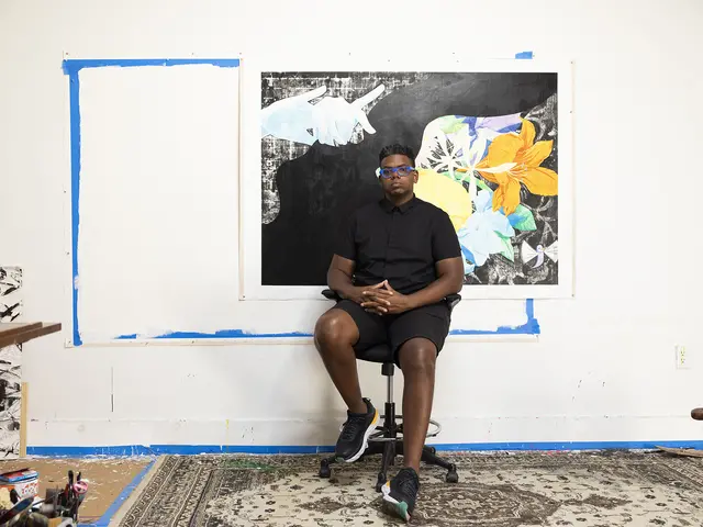 Mark sits on a black office chair in his studio. Blue painters tape frames the wall behind him, half covered by an in-progress work. He has dark skin and short black hair and wears a black button-up shirt, black shorts, and bright blue glasses.