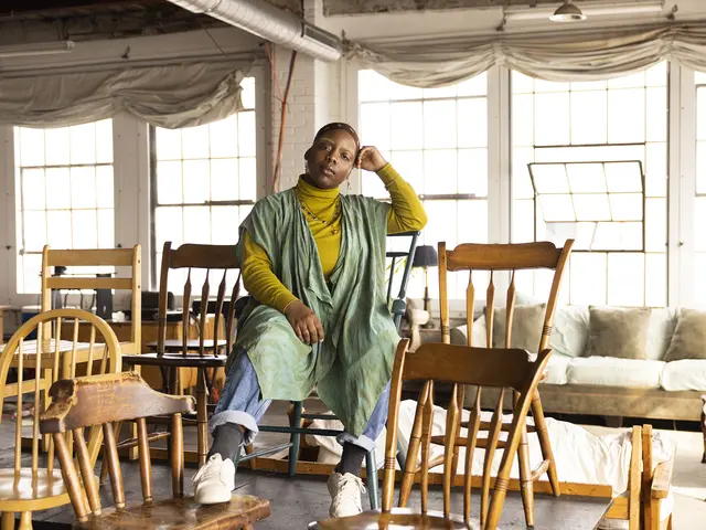 angel sits in an empty audience of mismatched wooden chairs in front of a row of bright windows. They have brown skin, blond eyebrows, and wear a kerchief on their head. They wear a yellow turtleneck sweater, green over-layer, jewelry, and jeans.