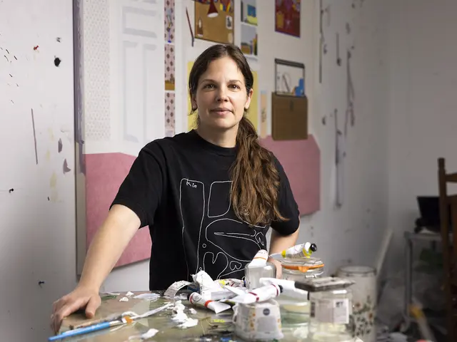 Becky Suss stands in her studio behind a work surface covered in empty jars and tubes of paint, a large in-process painting hanging behind her. She has light skin and long brown hair pulled into a ponytail over her shoulder. She wears a black graphic-T.