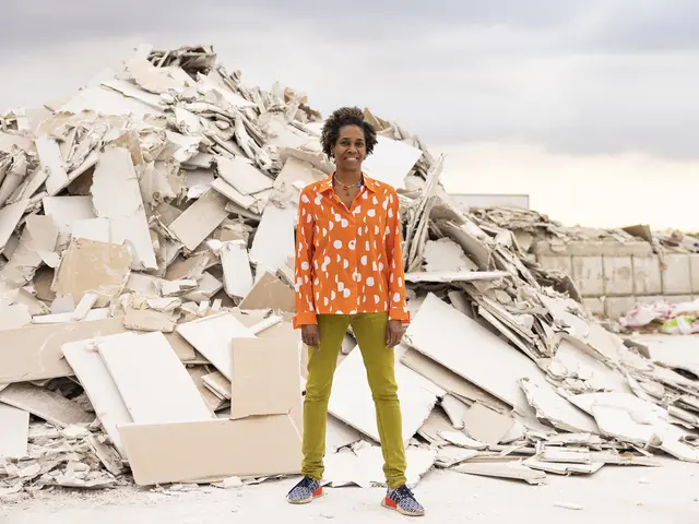 Karyn Olivier stands smiling in front of a large mound of broken drywall. She has brown skin and brown hair in tight curls with highlights. She is colorfully dressed in green pants, a patterned orange blouse, and black/white/orange patterned tennis shoes.